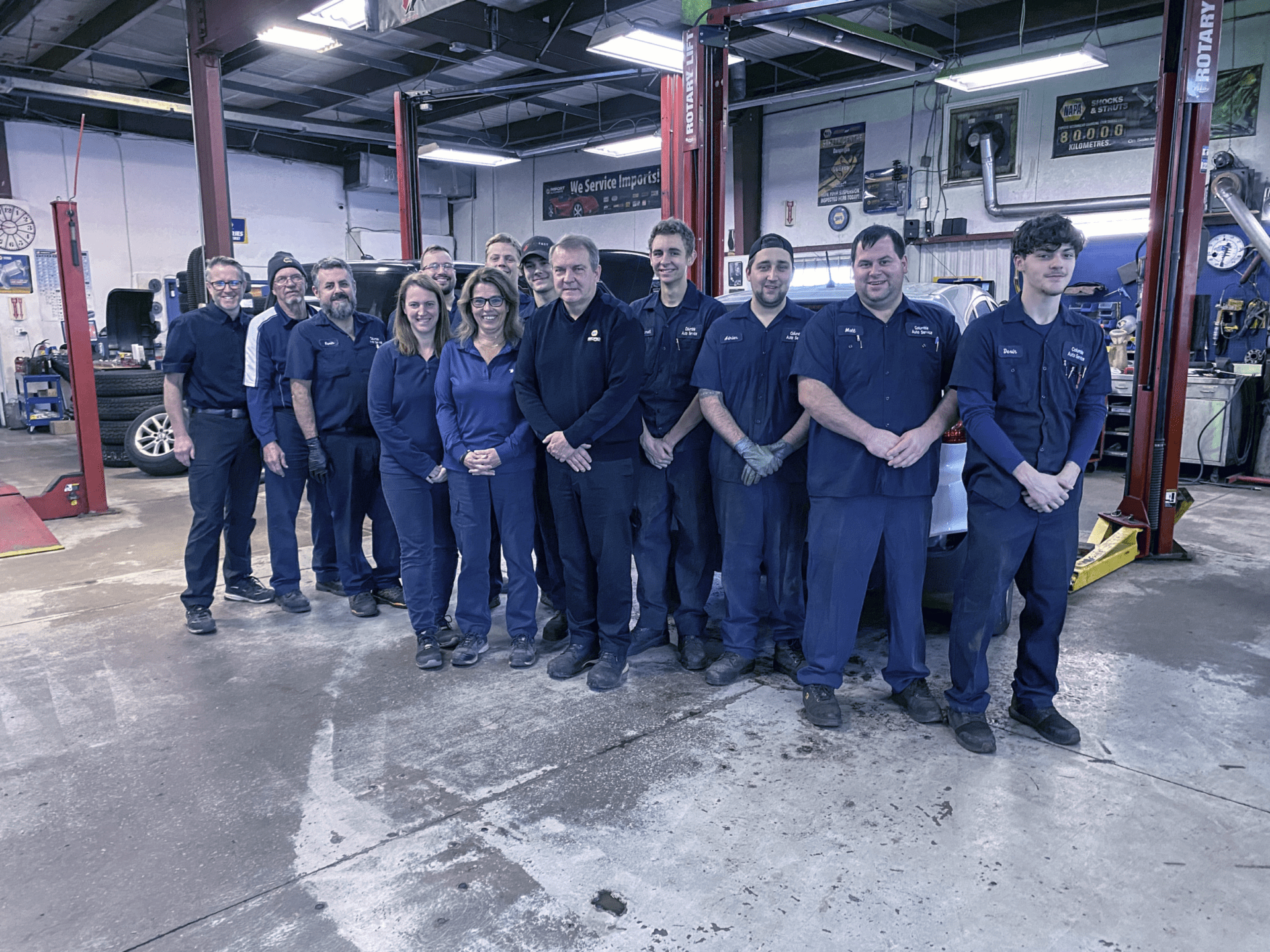 A group of people pose in a car repair workshop, wearing uniforms. They stand in front of vehicles and garage equipment, smiling at the camera.