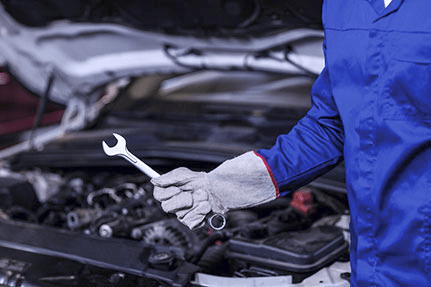 A person in a blue uniform with protective gloves holds a wrench while standing in front of an open car hood, suggesting auto repair or maintenance work.