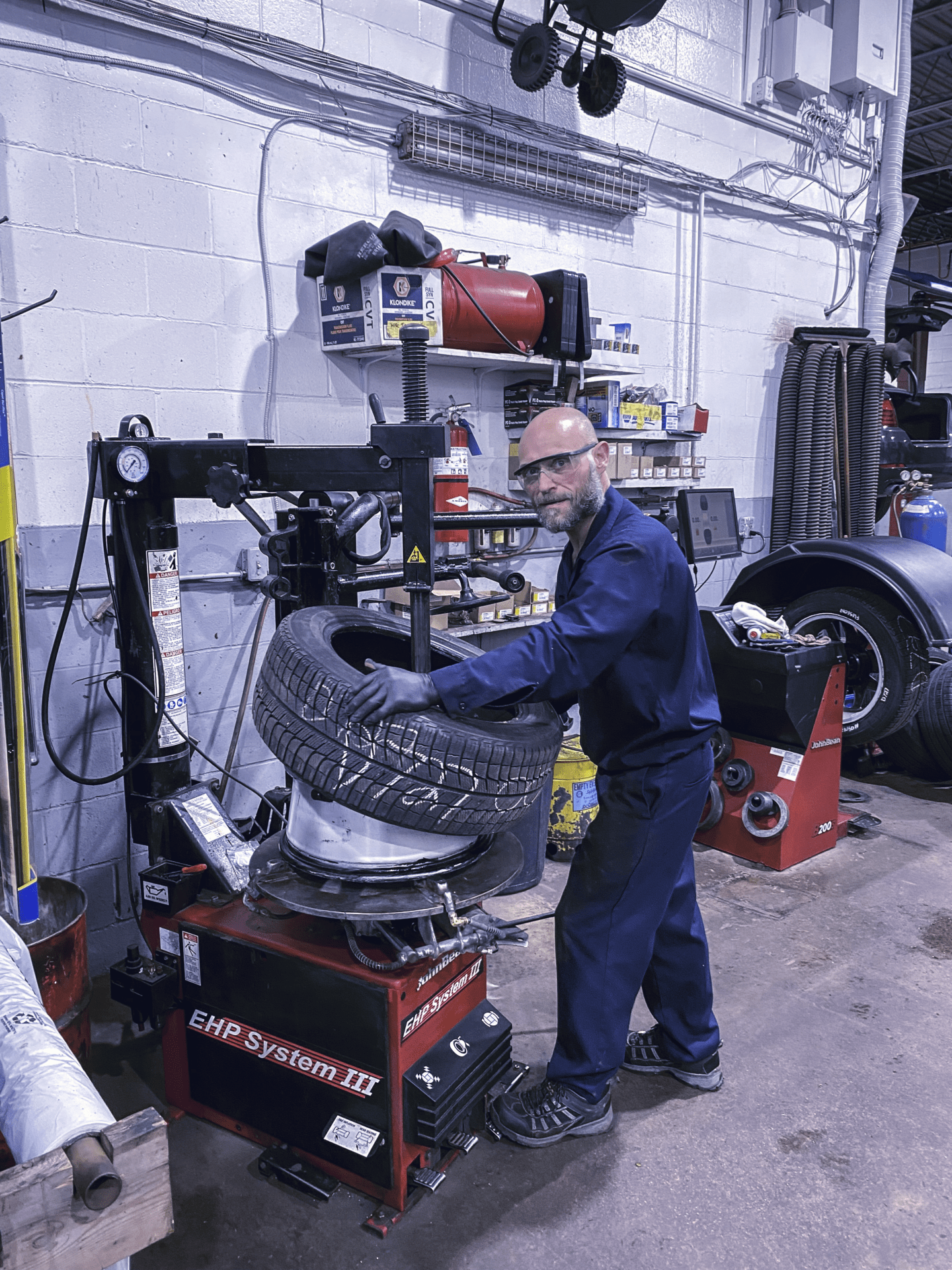 A person in a blue uniform is operating a tire changing machine in a well-equipped workshop. Tools, tires, and industrial equipment are visible.