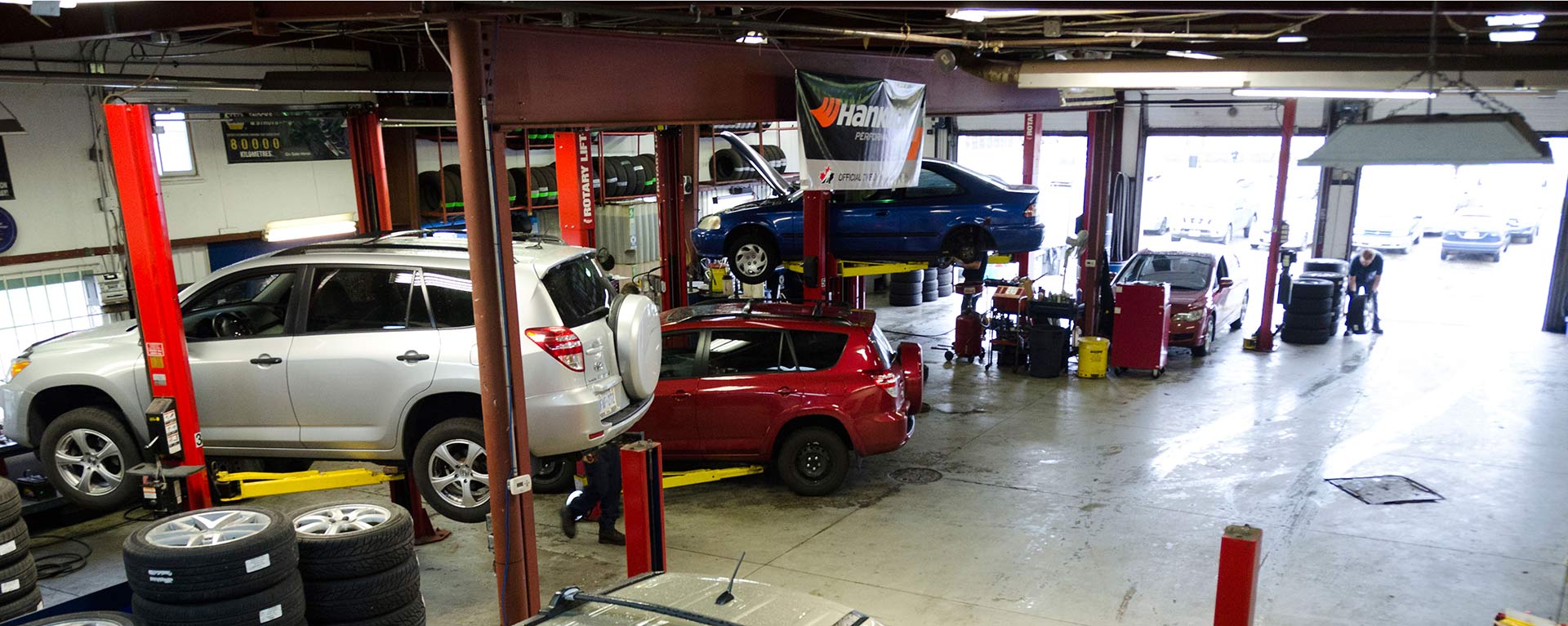 The image shows an active car repair garage with multiple vehicles lifted on hoists, a tire stack on the side, and at least one person working.
